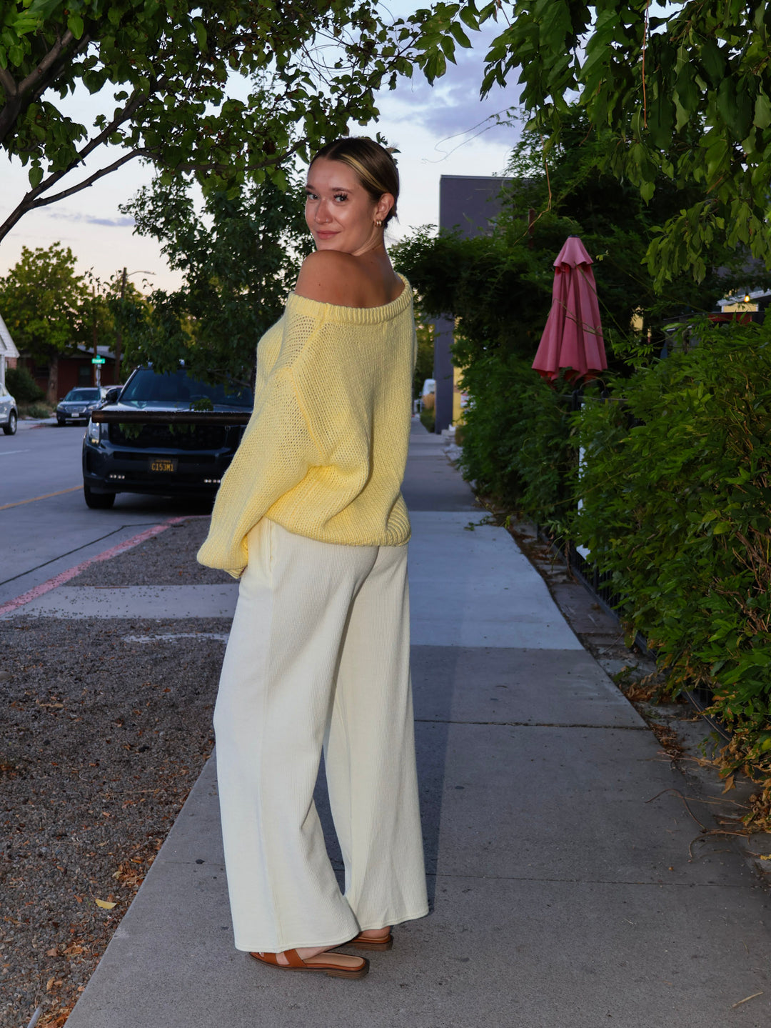Woman in a yellow sweater and white pants standing on a sidewalk with trees and a street in the background.
