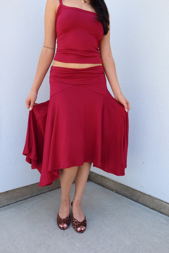 A woman wearing a matching two-piece outfit consisting of a sleeveless red crop top and a high-waisted red flared midi skirt, holding out the sides of the skirt to show its drape. She is wearing leopard print heels and standing against a light blue-gray stucco wall.