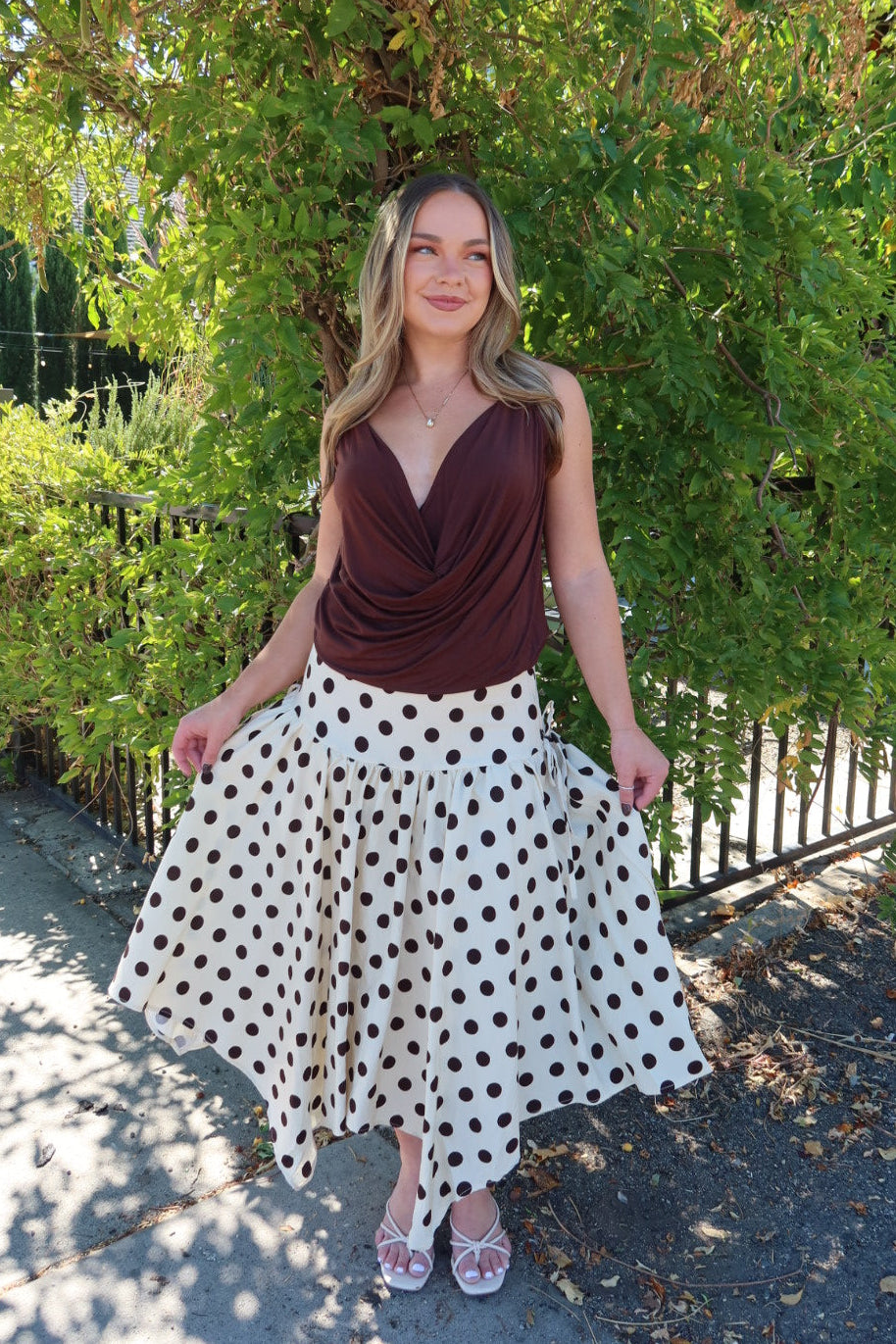 A woman stands outdoors in front of a leafy green tree, wearing a sleeveless dark brown draped top and a white midi skirt with brown polka dots. She is holding out the sides of the skirt to show its full flowy shape.