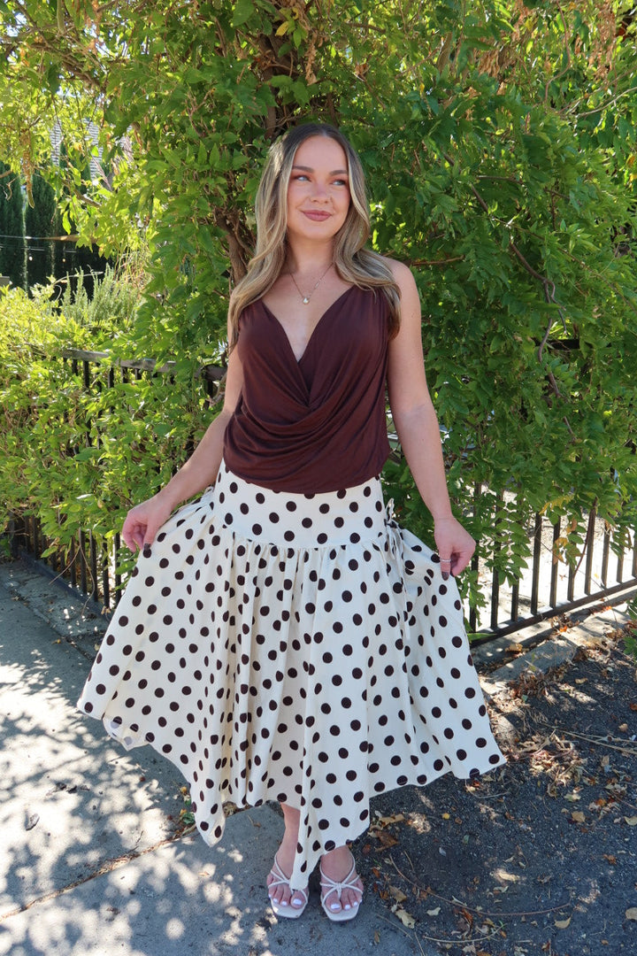 A woman stands outdoors in front of a leafy green tree, wearing a sleeveless dark brown draped top and a white midi skirt with brown polka dots. She is holding out the sides of the skirt to show its full flowy shape.