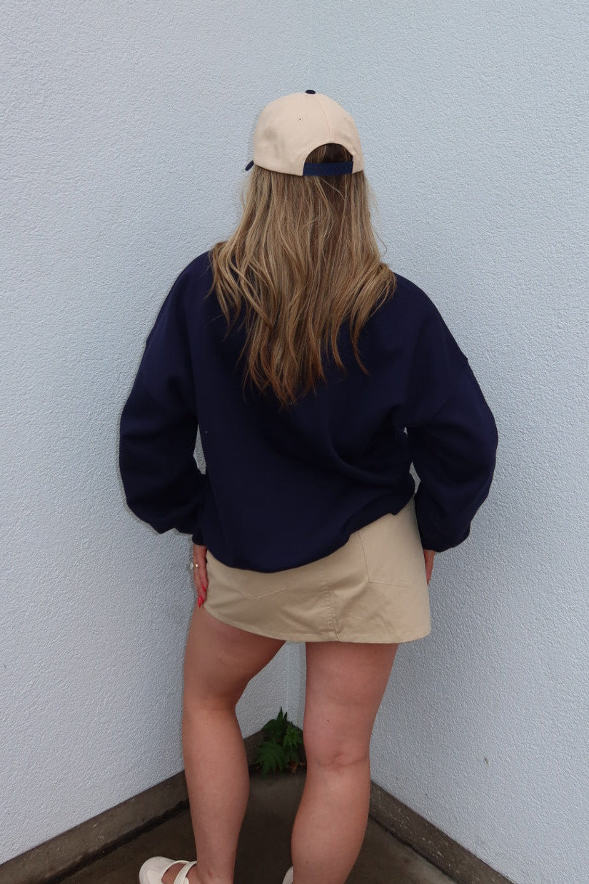 A person viewed from behind wearing a navy blue sweatshirt, a beige mini skirt, a beige baseball cap, and white sandals, standing in the corner of a white wall.