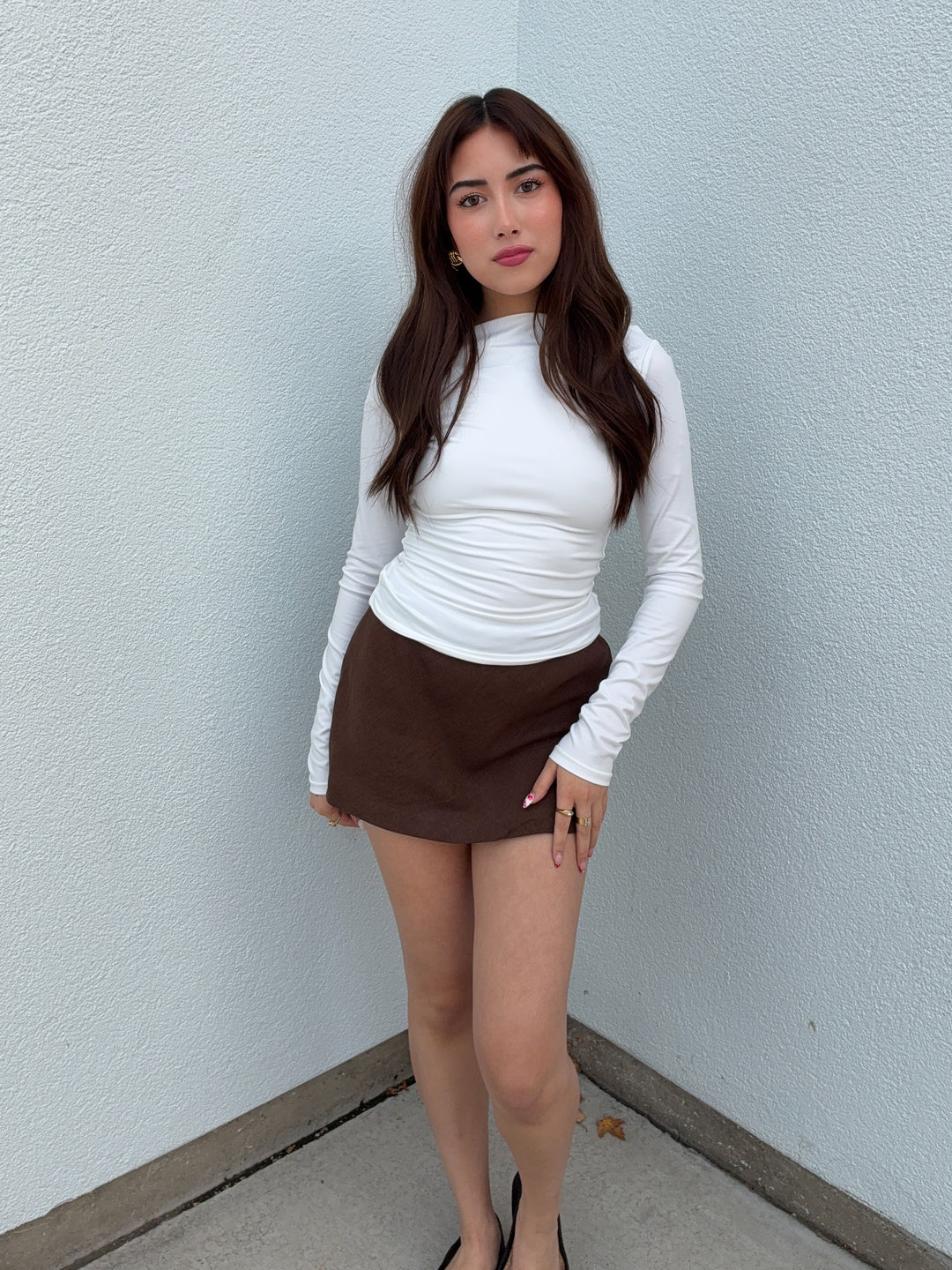 Woman wearing a white long-sleeve top and brown skirt against a light blue wall.