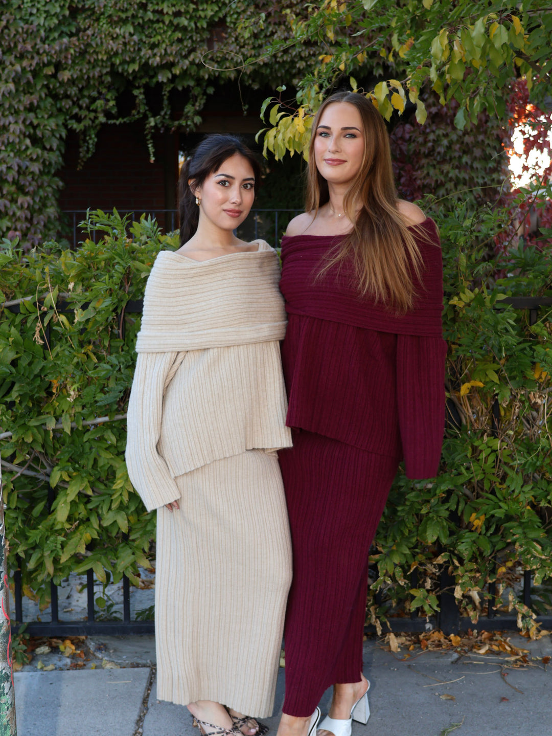 Two women standing outdoors wearing matching knit skirt sets with greenery in the background