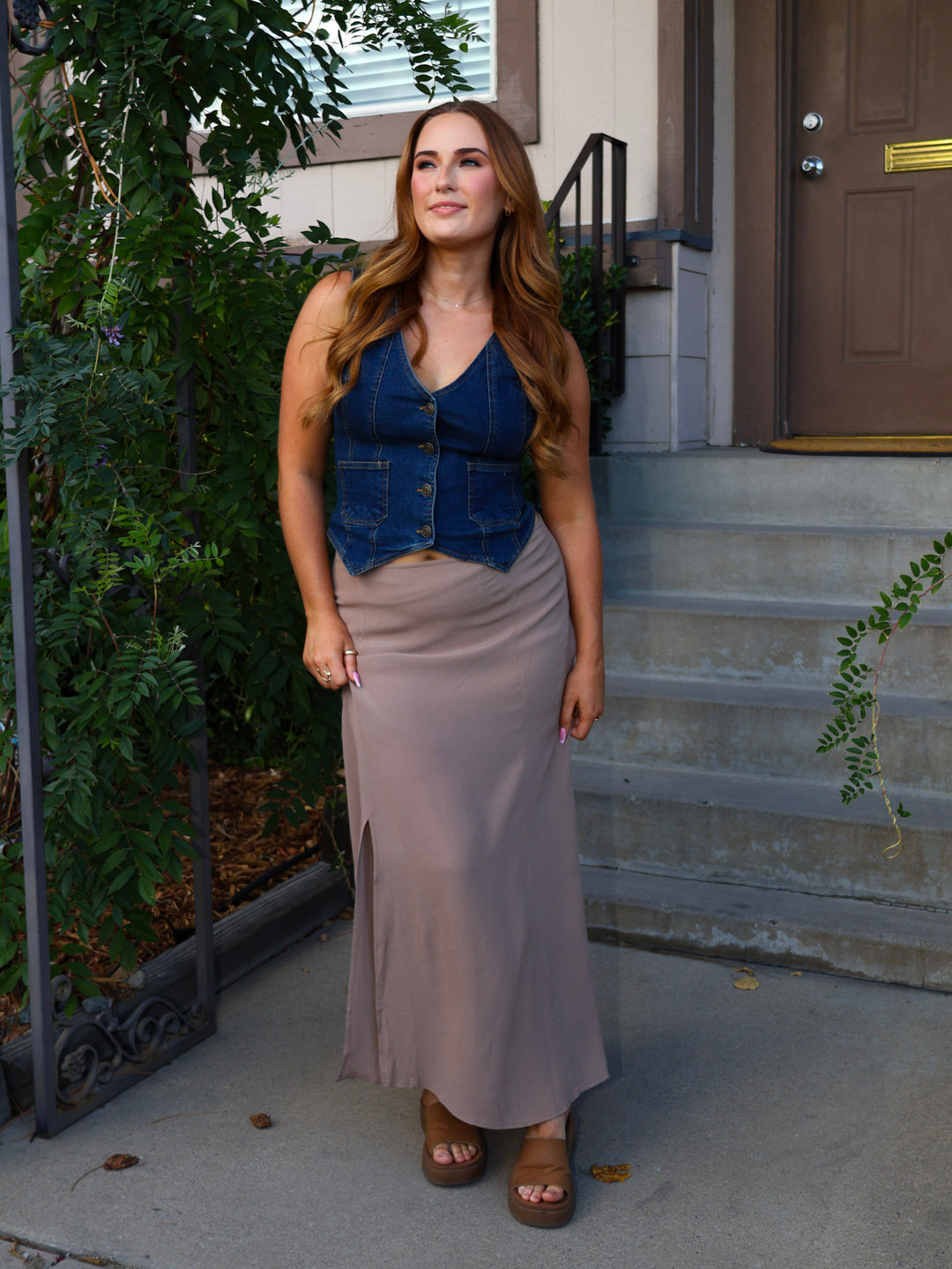 Woman in a blue top and gray skirt standing on a sidewalk with plants and a door in the background