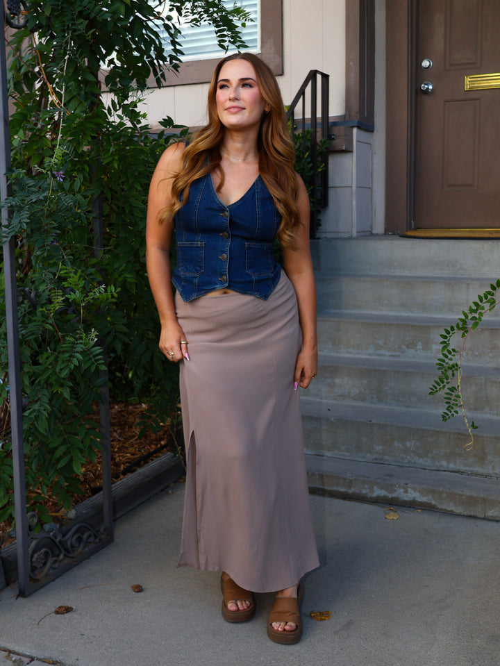 Woman in a blue top and gray skirt standing on a sidewalk with plants and a door in the background