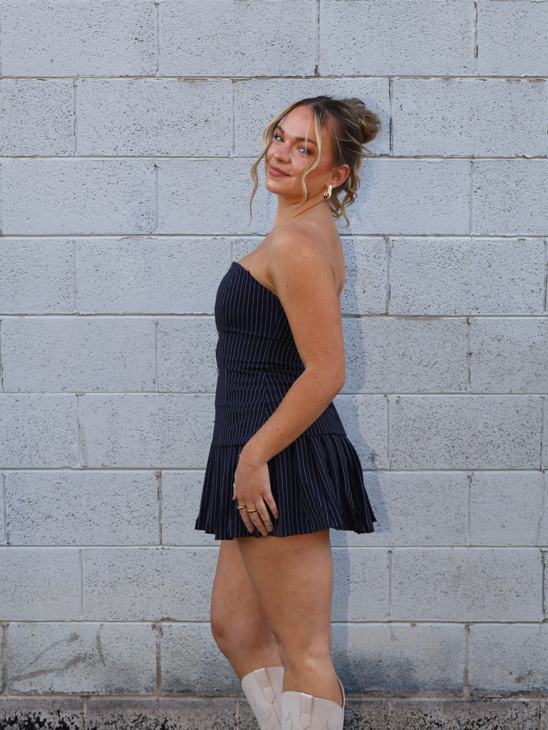 Woman in a navy pleated dress standing against a gray brick wall.