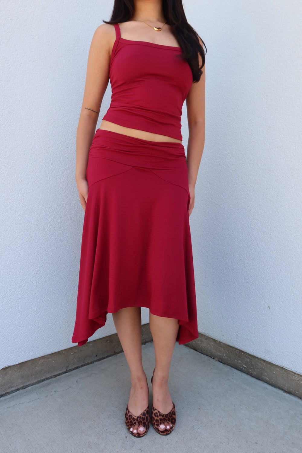 A woman modeling a matching two-piece red outfit consisting of a sleeveless crop top and a draped high-waisted midi skirt, paired with leopard print heels, standing against a light gray wall.