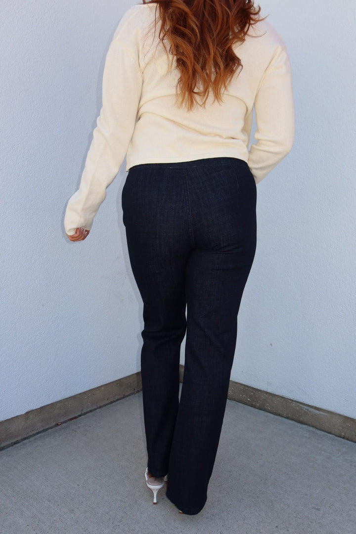 A woman viewed from behind wearing dark navy blue jeans and a cream-colored long-sleeve top, standing in a corner against a light blue wall.