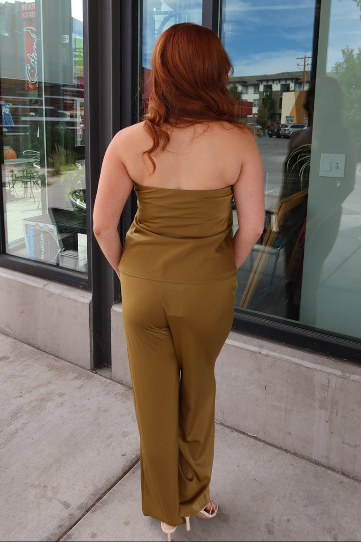 A woman with red hair is seen from behind wearing a strapless olive green jumpsuit while standing on a sidewalk in front of a storefront with large windows.