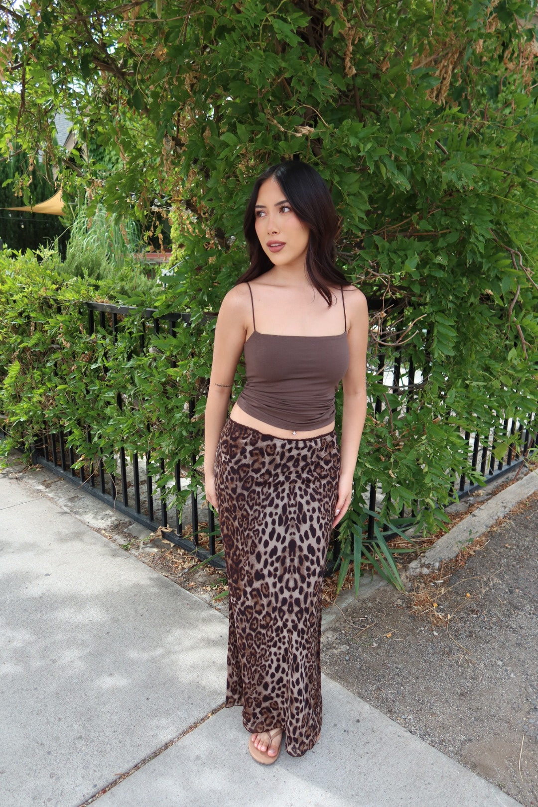 A woman stands outdoors on a sidewalk in front of green bushes and a black metal fence, wearing a brown ribbed spaghetti-strap crop top and a brown leopard-print maxi skirt.