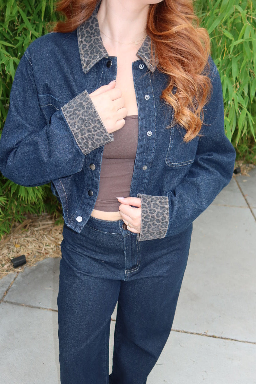 A woman wearing a dark blue denim jacket and matching wide-leg jeans with leopard-print collar and cuff accents, paired with a brown top and sandals, standing outdoors on a concrete patio with green foliage in the background.
