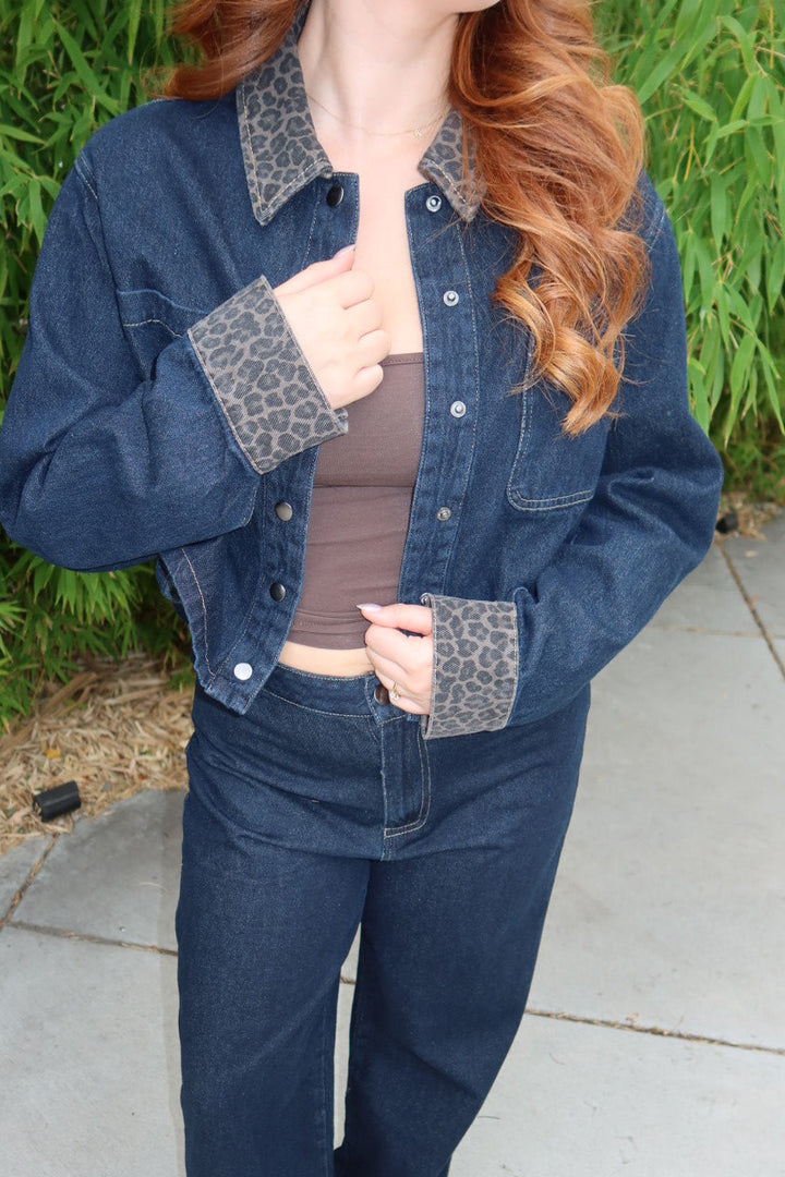 A woman wearing a dark blue denim jacket and matching wide-leg jeans with leopard-print collar and cuff accents, paired with a brown top and sandals, standing outdoors on a concrete patio with green foliage in the background.