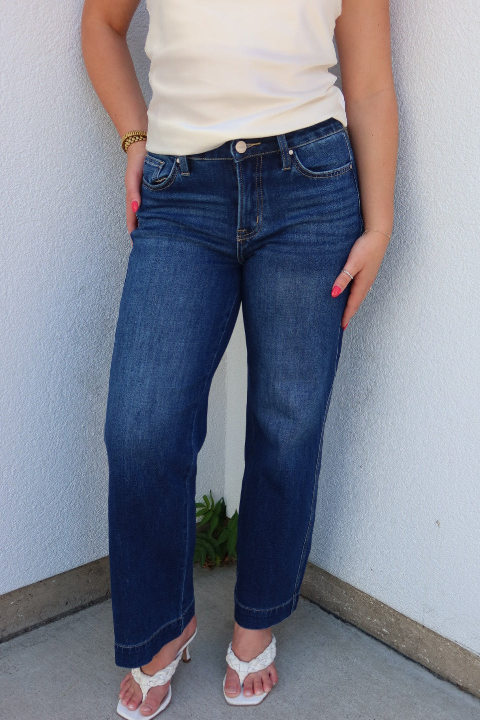 A person wearing dark blue straight-leg jeans, a cream sleeveless top, and white heeled sandals, standing against a light gray textured wall.