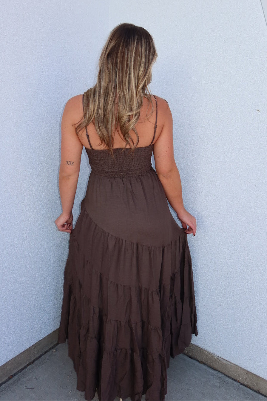 A woman viewed from behind wearing a brown spaghetti-strap maxi dress with a tiered skirt, standing against a white wall.