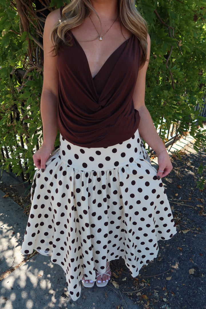 A woman wearing a dark brown draped tank top and a white skirt with brown polka dots, standing outdoors in front of green foliage.