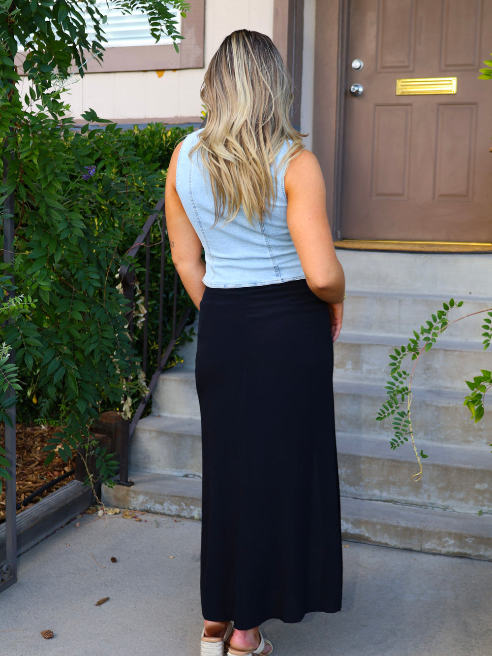Woman standing outside a house wearing a light blue sleeveless top and black skirt.
