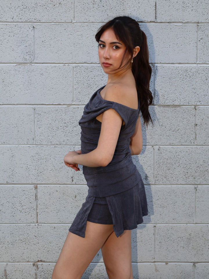 Woman wearing a gray off-shoulder top and skirt against a gray brick wall.