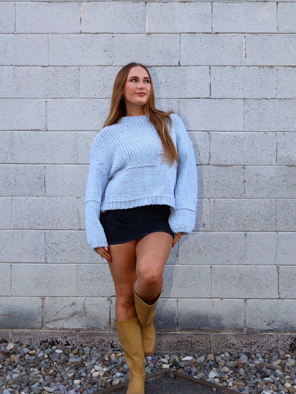 Woman wearing a light blue sweater and beige knee-high boots standing against a gray brick wall.