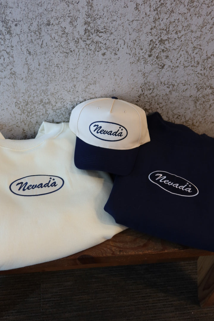 Three folded sweatshirts and one baseball cap with 'Nevada' embroidered in an oval logo, arranged on a wooden surface against a textured gray wall.