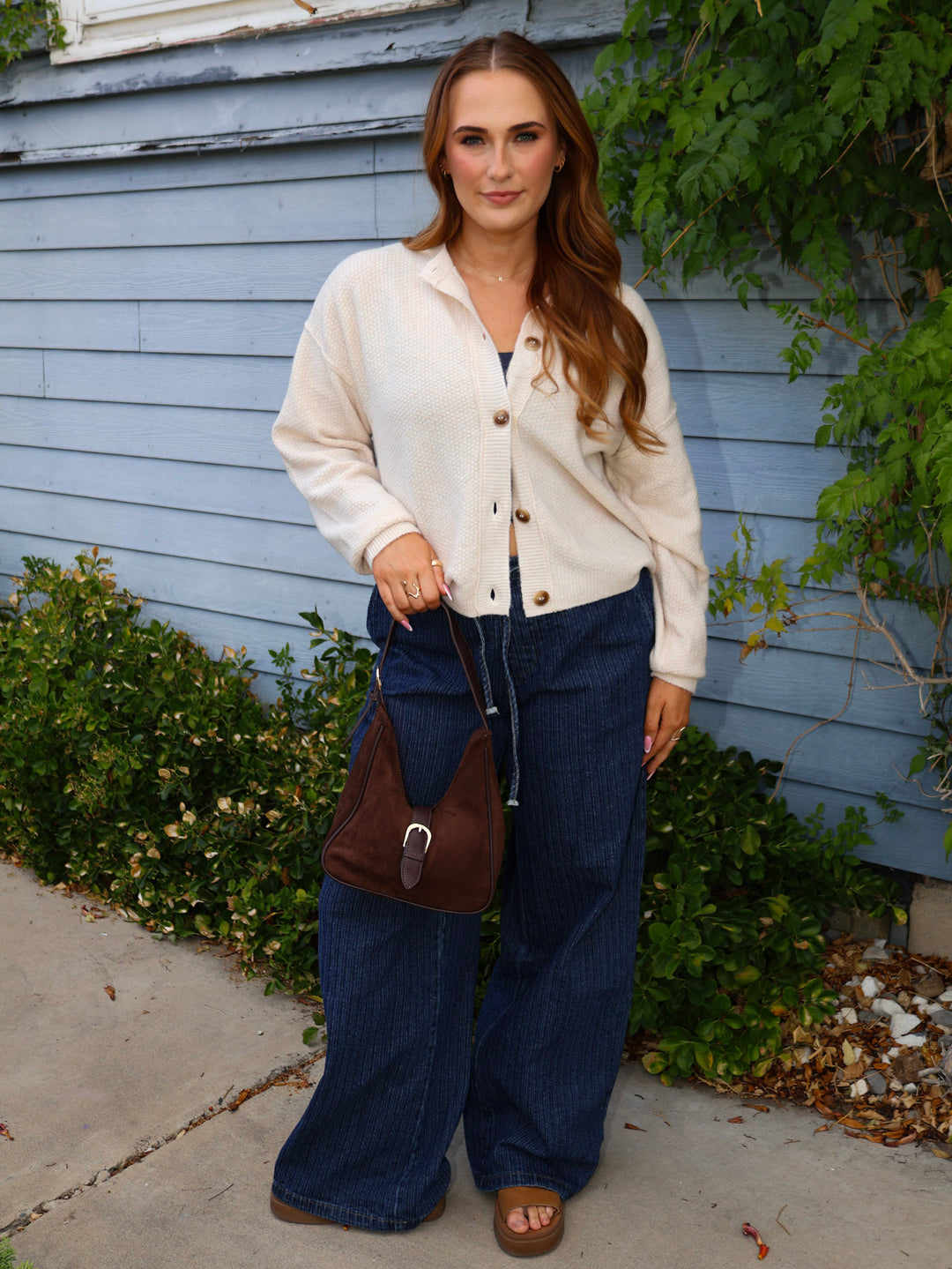 Woman wearing a cream cardigan, blue jeans, and brown sandals standing in front of a light blue house.