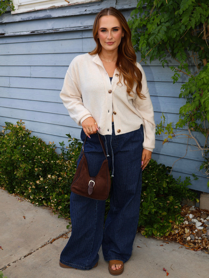 Woman wearing a cream cardigan, blue jeans, and brown sandals standing in front of a light blue house.