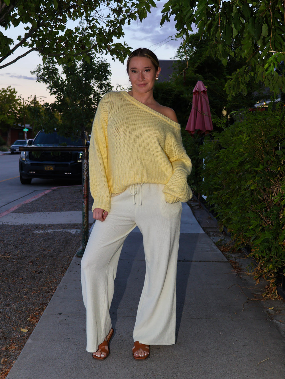Woman wearing a yellow sweater and white pants standing outdoors on a street.