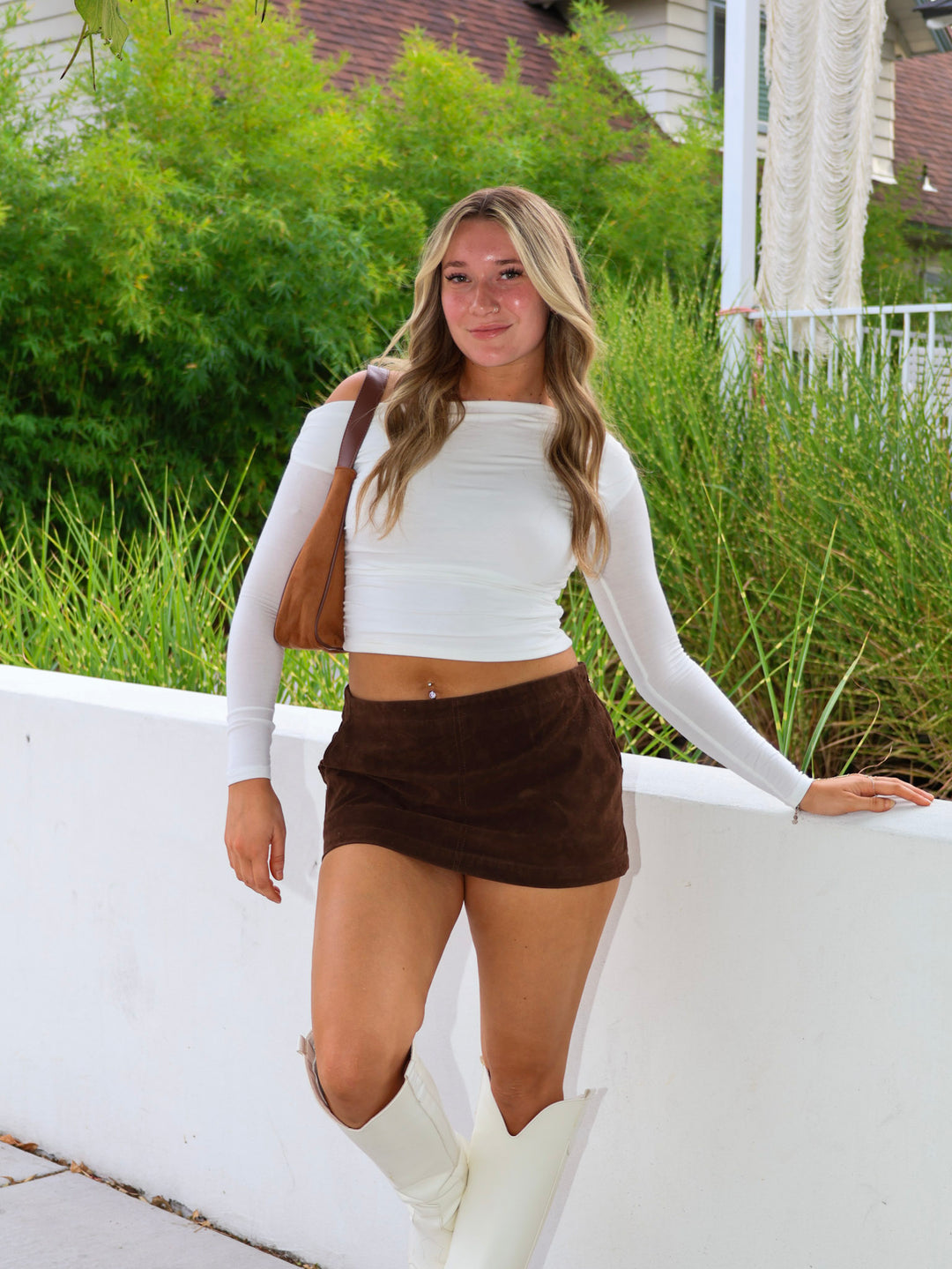 Woman in white long-sleeve top and brown shorts standing outdoors with greenery in the background