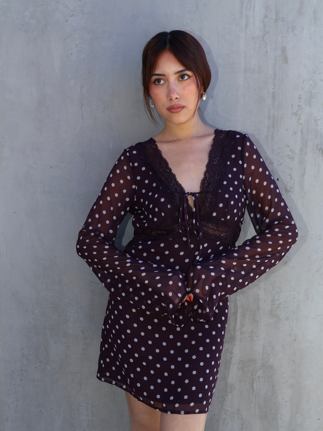 Woman wearing a sheer brown dress with white polka dots against a gray wall.