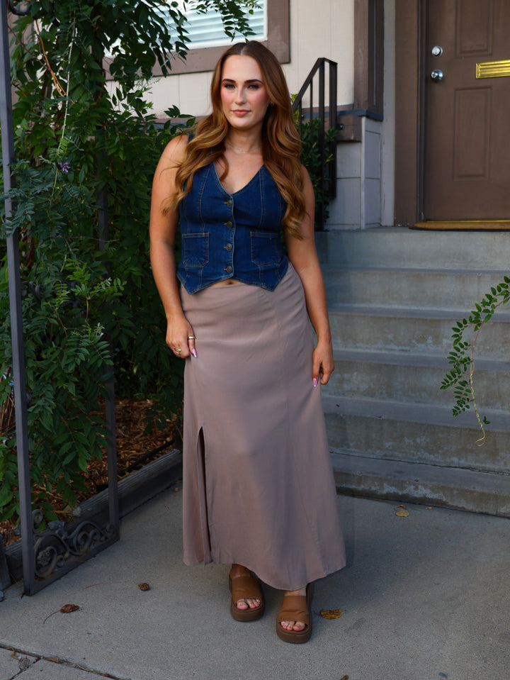 Woman standing on a sidewalk wearing a denim vest over a long skirt.