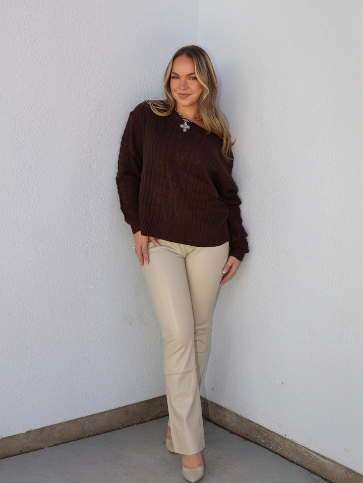 Woman wearing a brown sweater and beige pants against a white wall.