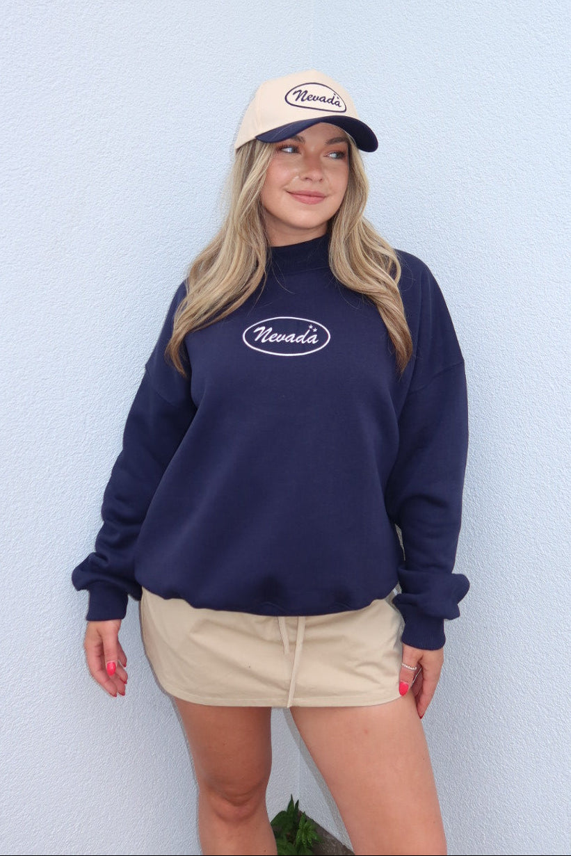 A woman wearing a navy blue sweatshirt with a white 'Nevada' logo, a beige baseball cap with 'Nevada' on the front, and a beige mini skirt, posing against a light blue textured wall.