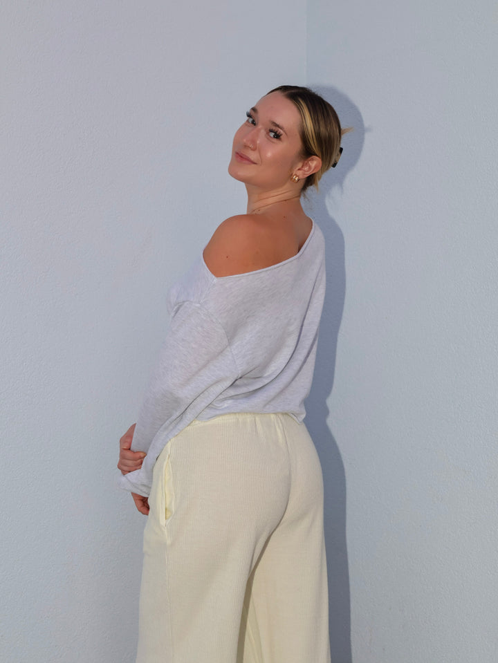 Woman wearing a light gray off-shoulder top and beige pants against a plain background
