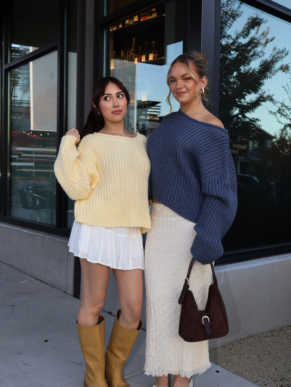 Two women standing on a sidewalk, one in a yellow sweater and white skirt, the other in a blue sweater and white skirt.