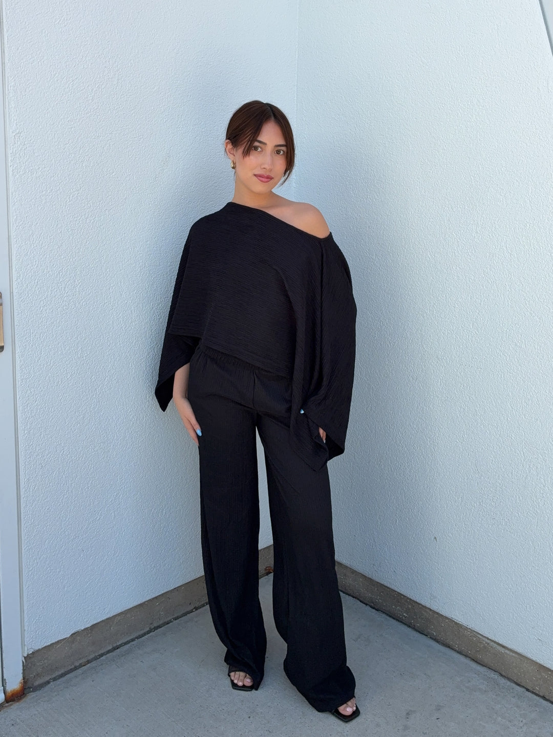 Woman wearing a black off-shoulder top and wide-leg pants against a light blue wall.