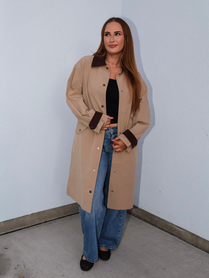 Woman wearing a beige trench coat and blue jeans against a white wall.