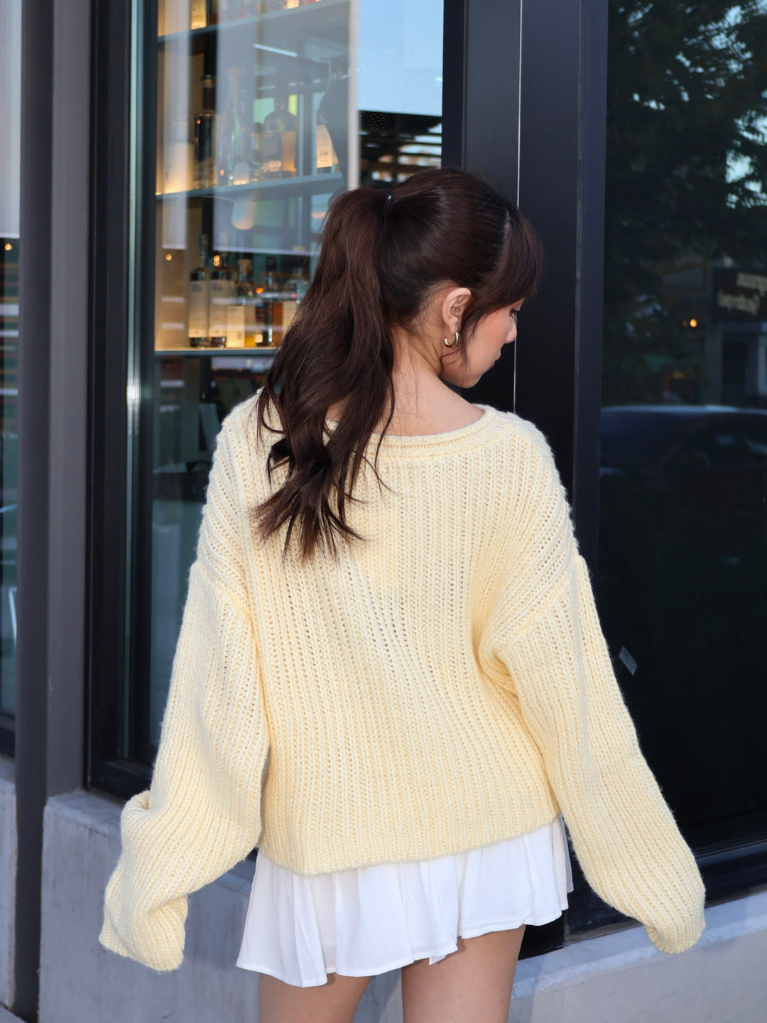 Woman wearing a light yellow sweater and white skirt standing outside a building.