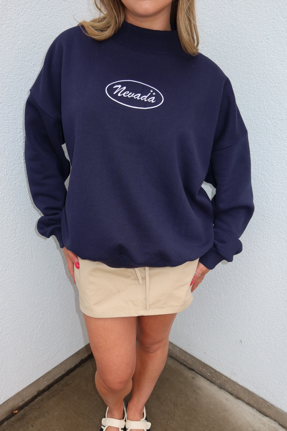 A person wearing an oversized navy blue sweatshirt with a white 'Nevada' logo on the chest, paired with a beige mini skirt and white Mary Jane shoes, standing in a corner against a light gray wall.