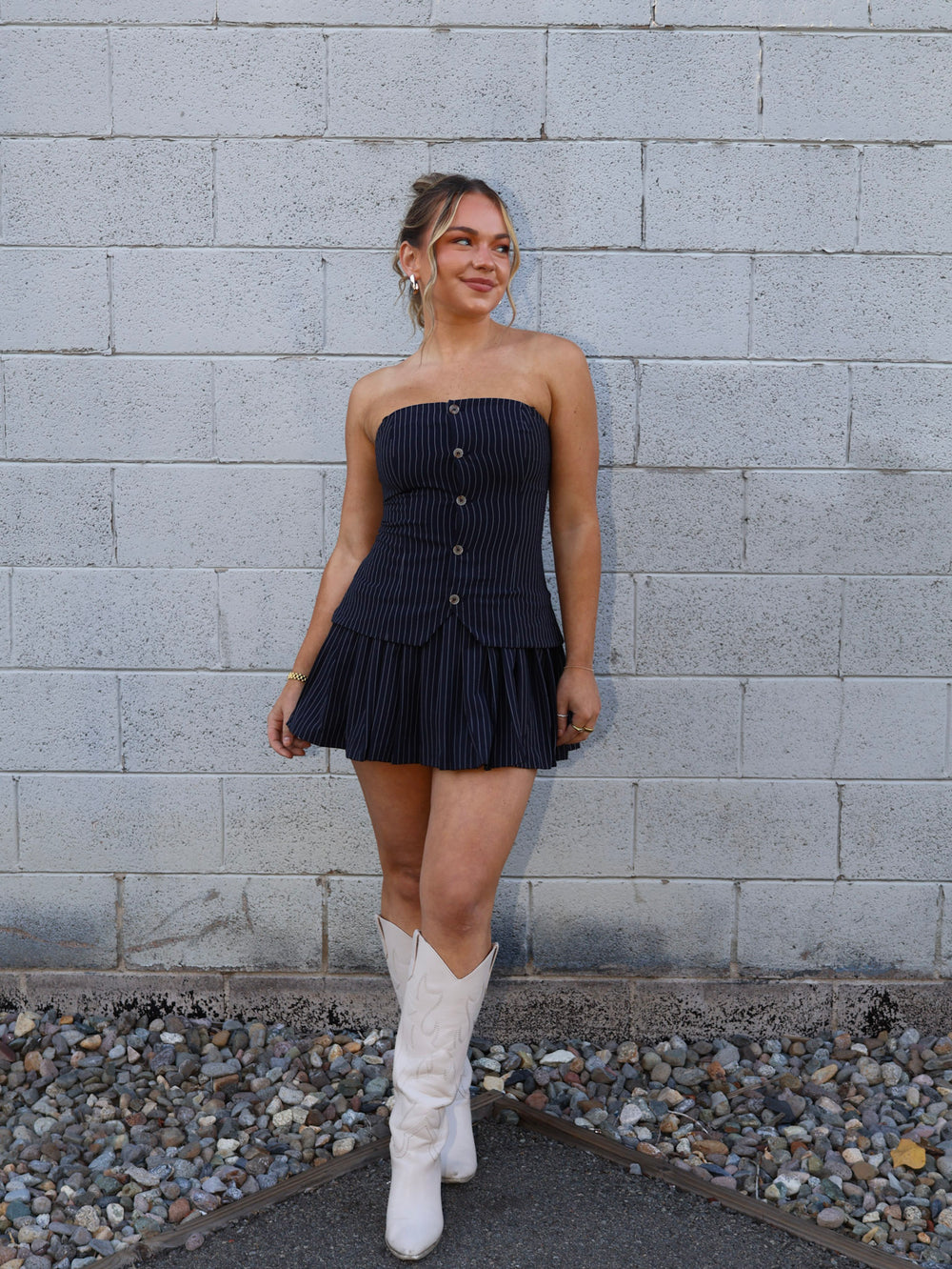 Woman in a strapless navy dress and white boots standing against a gray brick wall.