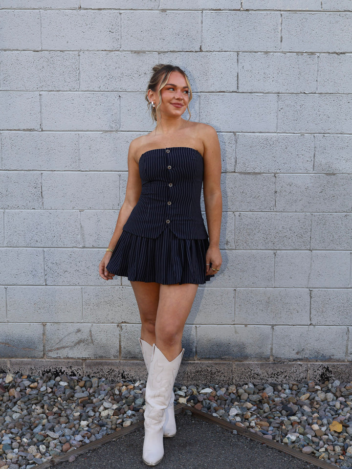 Woman in a strapless navy dress and white boots standing against a gray brick wall.