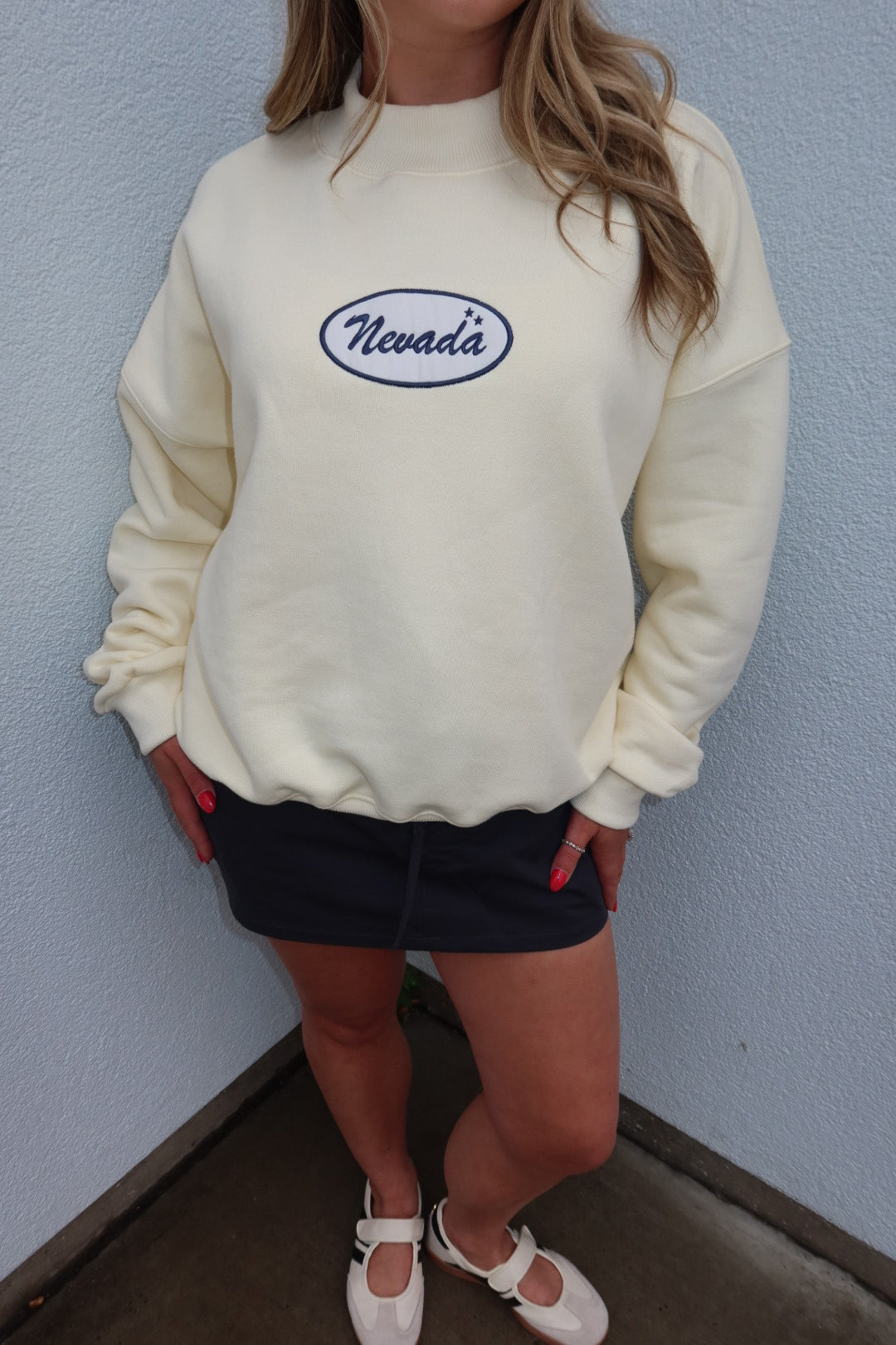 A person wearing a cream-colored crewneck sweatshirt with an embroidered oval Nevada logo on the chest, paired with a dark skirt and white shoes, standing against a light blue textured wall.