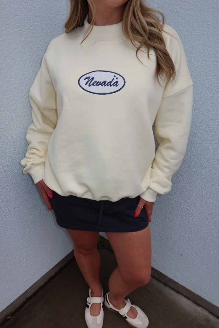 A person wearing a cream-colored crewneck sweatshirt with an embroidered oval Nevada logo on the chest, paired with a dark skirt and white shoes, standing against a light blue textured wall.