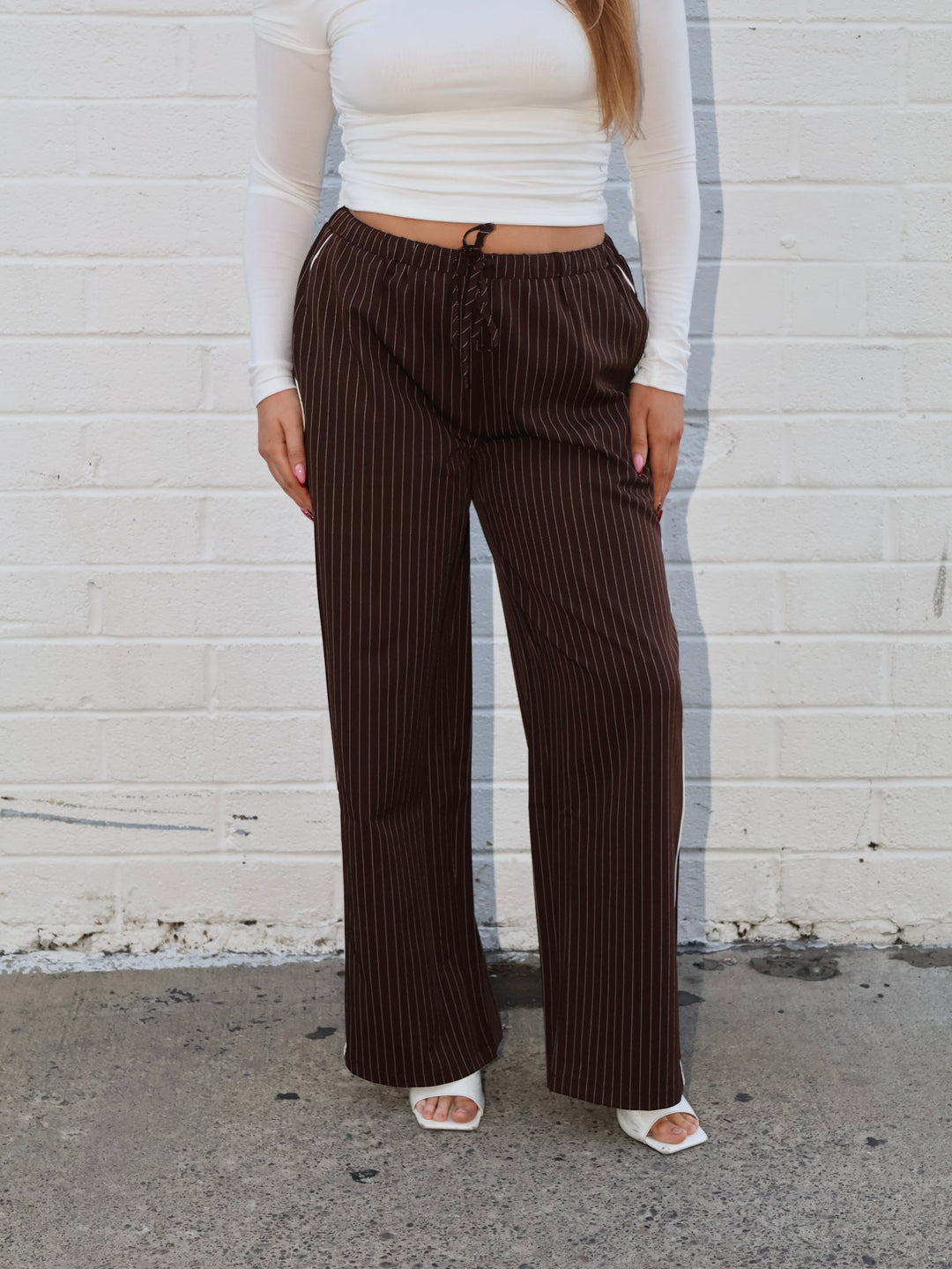 Person wearing brown pinstripe pants and a white top against a white brick wall.