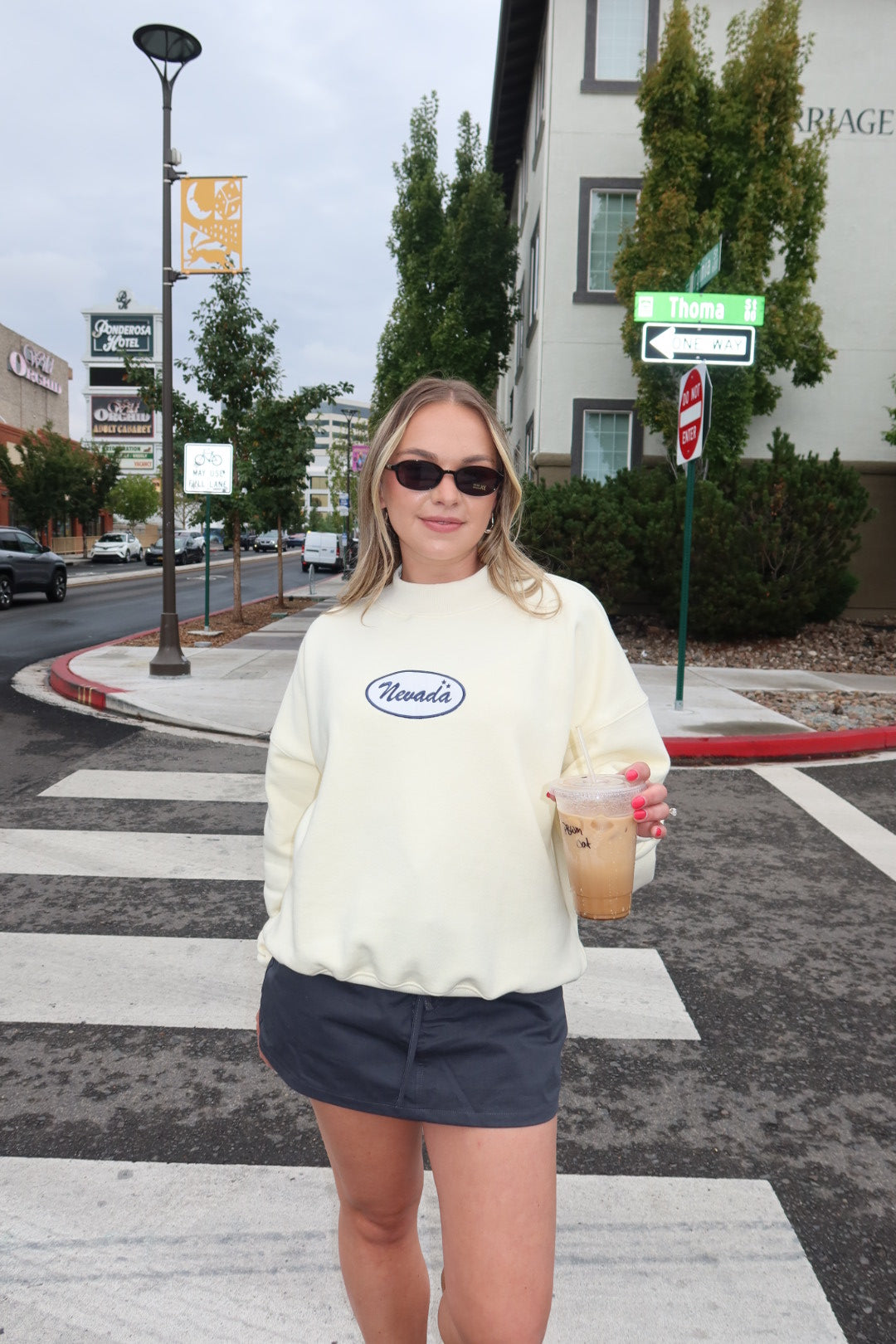 A woman standing on a city crosswalk wearing a cream-colored crewneck sweatshirt with a blue oval 'Nevada' logo on the chest, paired with a dark gray mini skirt and holding an iced coffee.