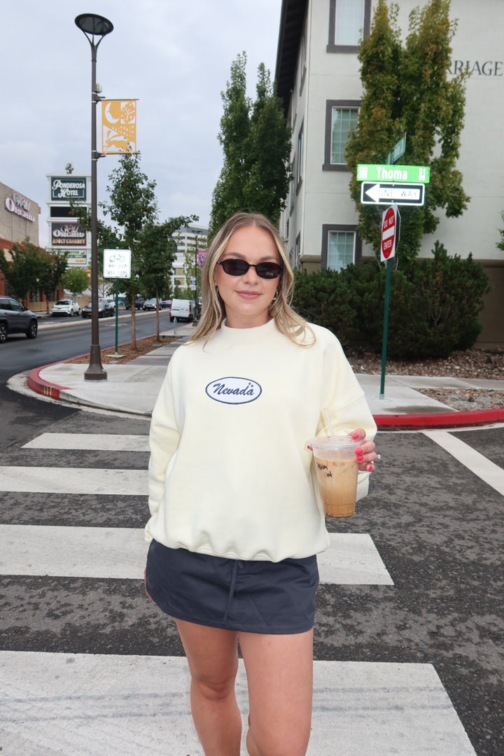 A woman standing on a city crosswalk wearing a cream-colored crewneck sweatshirt with a blue oval 'Nevada' logo on the chest, paired with a dark gray mini skirt and holding an iced coffee.