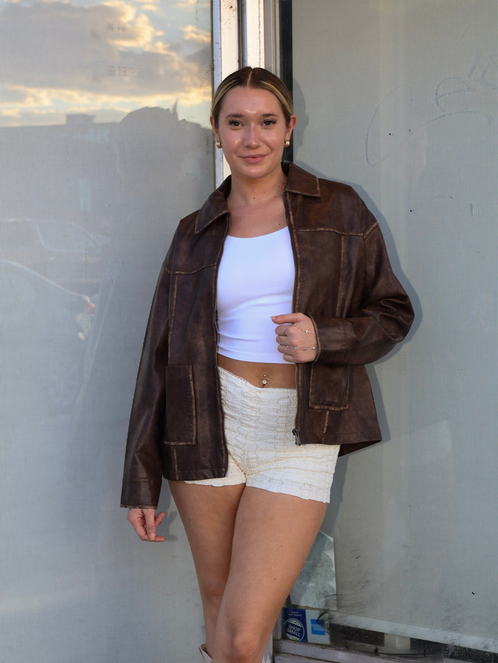 Woman wearing a brown leather jacket, white top, and white shorts standing against a plain wall.