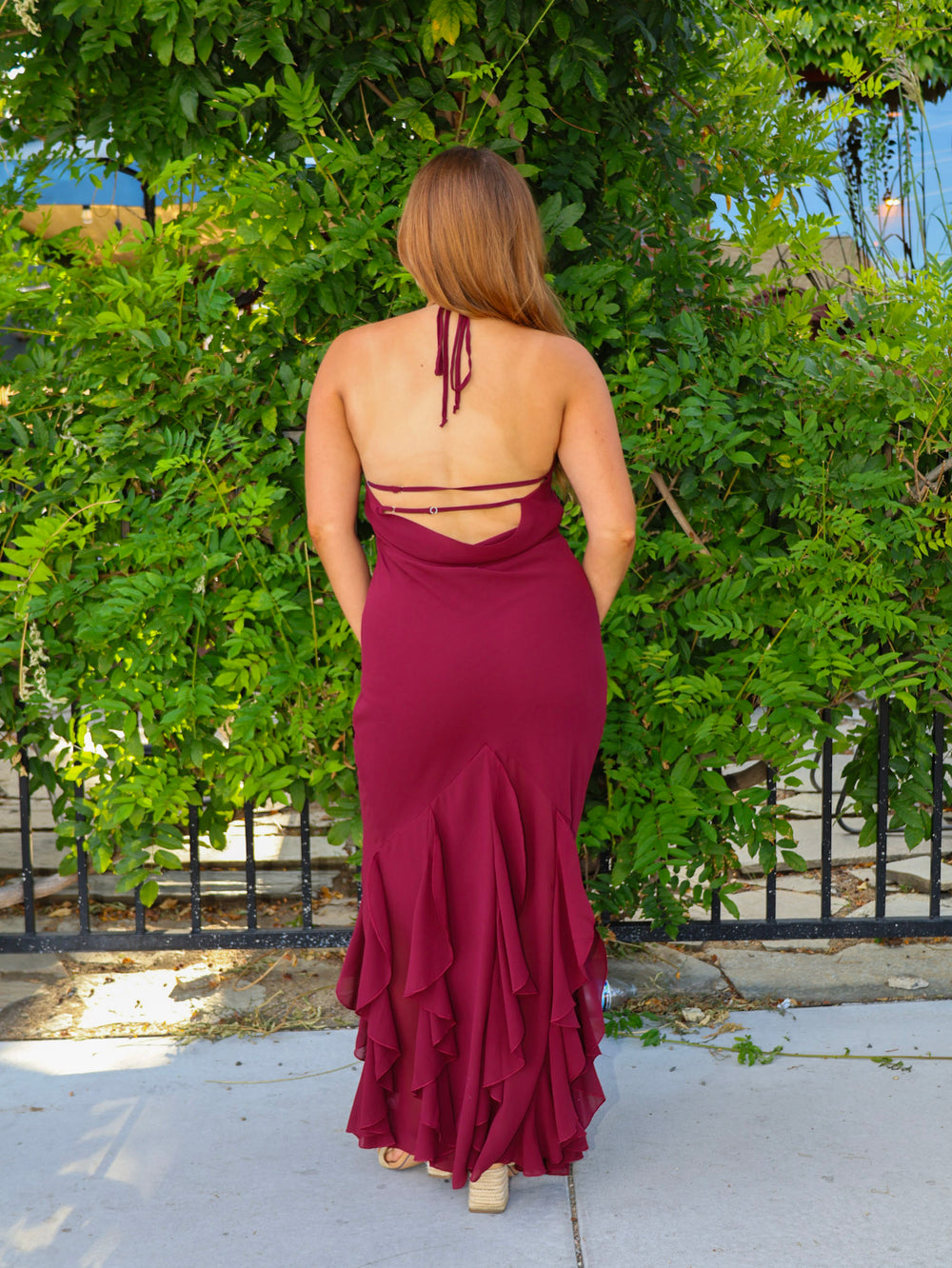 Woman wearing a burgundy dress with cut-out back and ruffled skirt, standing in front of green foliage.