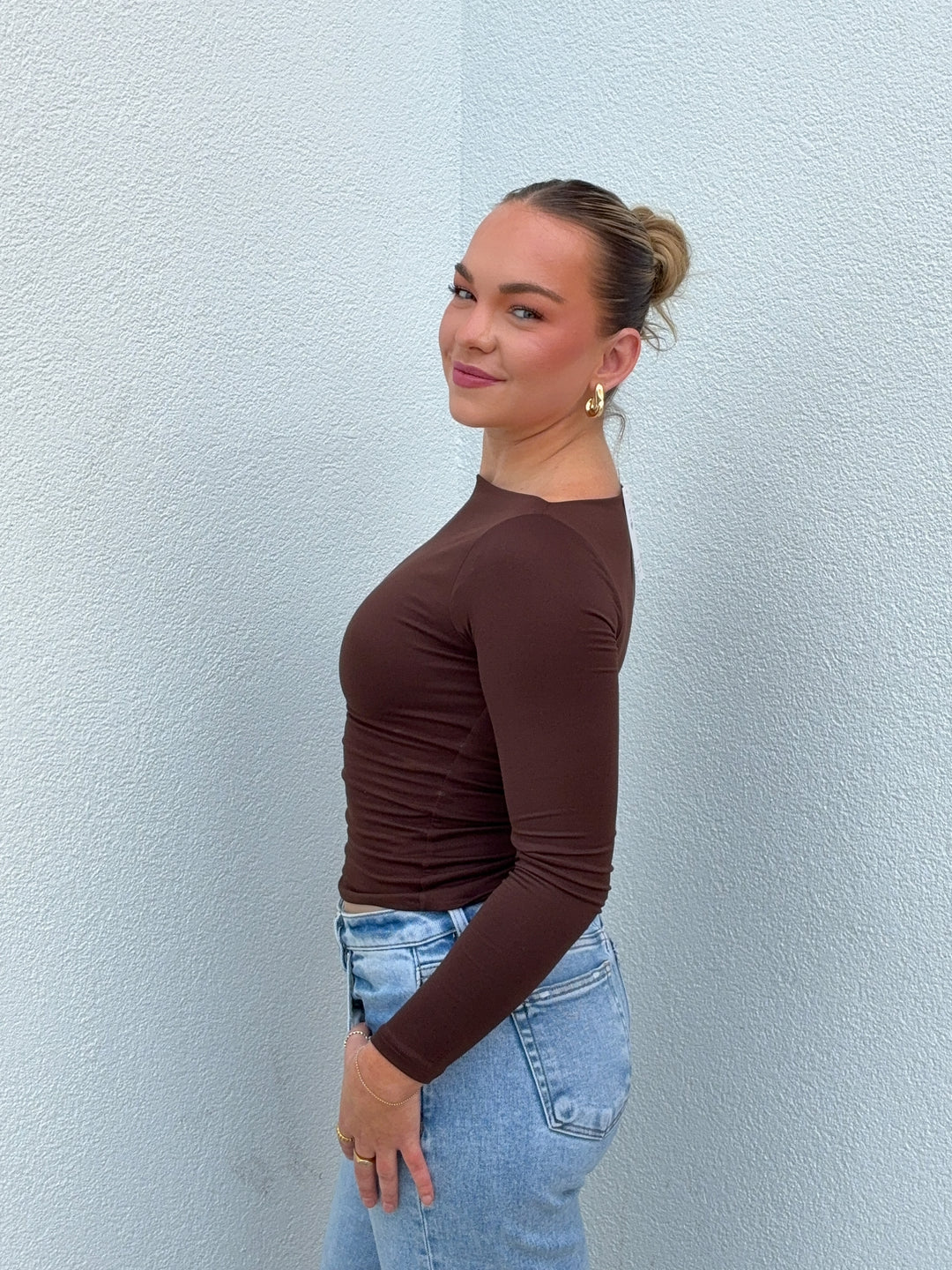 Woman wearing a brown long-sleeve top and blue jeans against a light blue wall.
