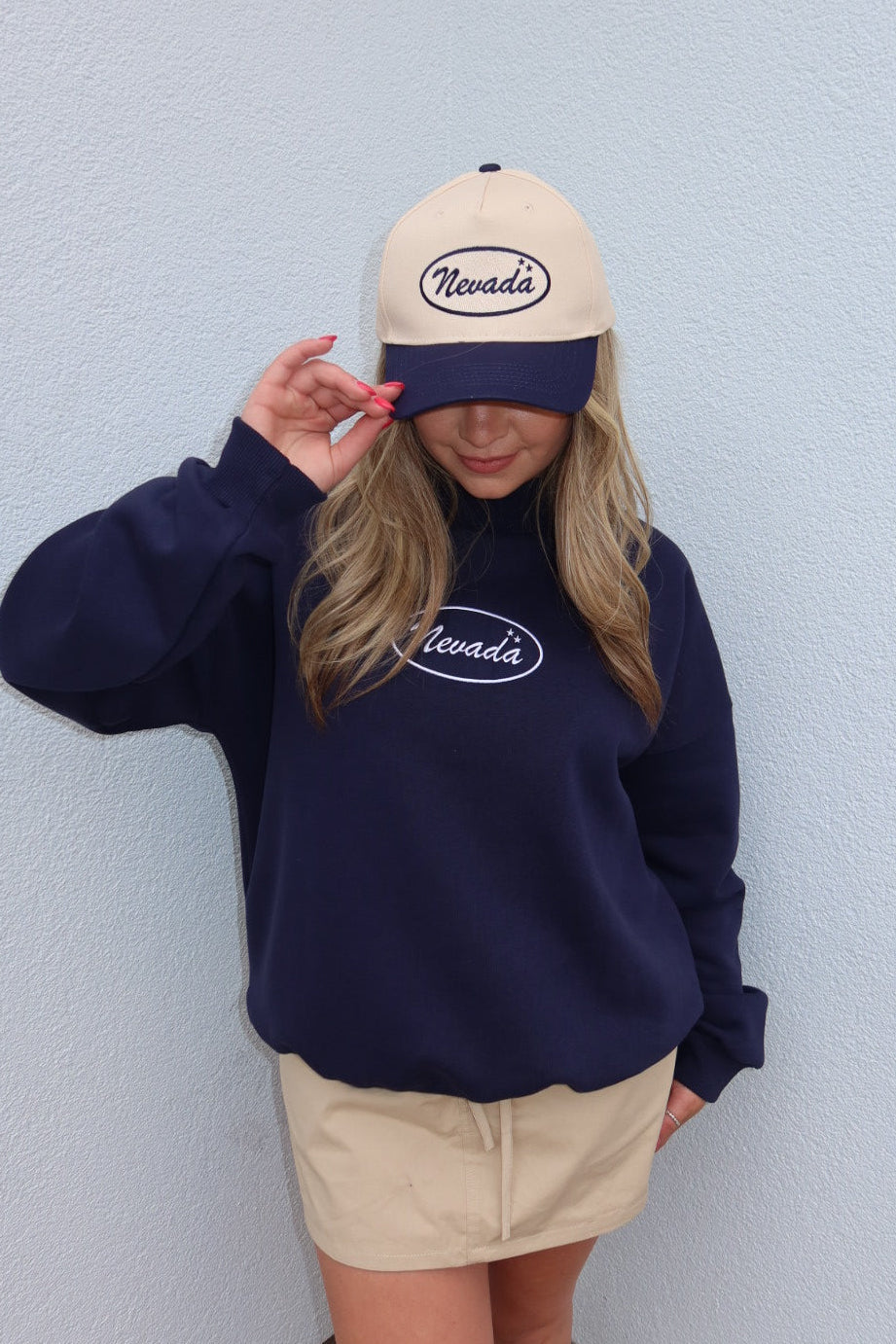 A woman wearing a navy blue sweatshirt and beige shorts, with a matching beige and navy baseball cap featuring the word 'Nevada' in an oval logo. She is posing against a light blue textured wall.