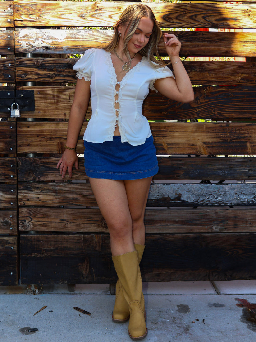 Woman in white blouse, blue skirt, and yellow boots standing against a wooden wall.