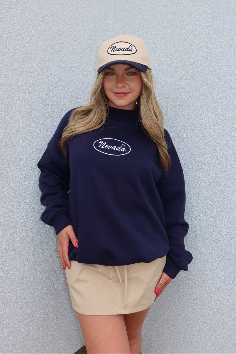 A woman wearing a navy blue sweatshirt with a white oval 'Nevada' logo, a beige baseball cap with 'Nevada' in script, and a beige mini skirt, standing against a light gray textured wall.